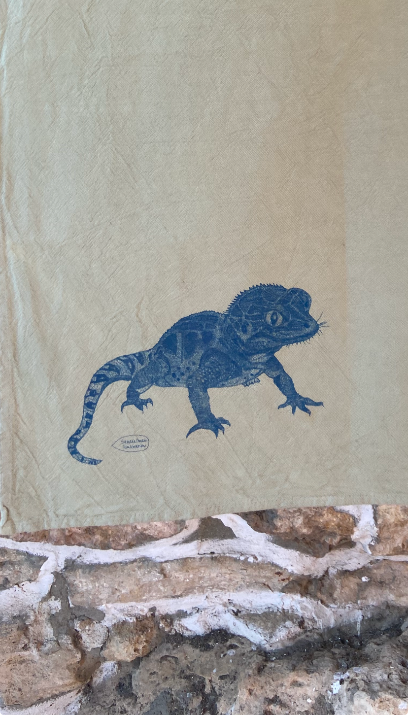 A beige fabric tapestry or wall hanging printed with a blue gecko design, shown draped over a rustic stone wall.